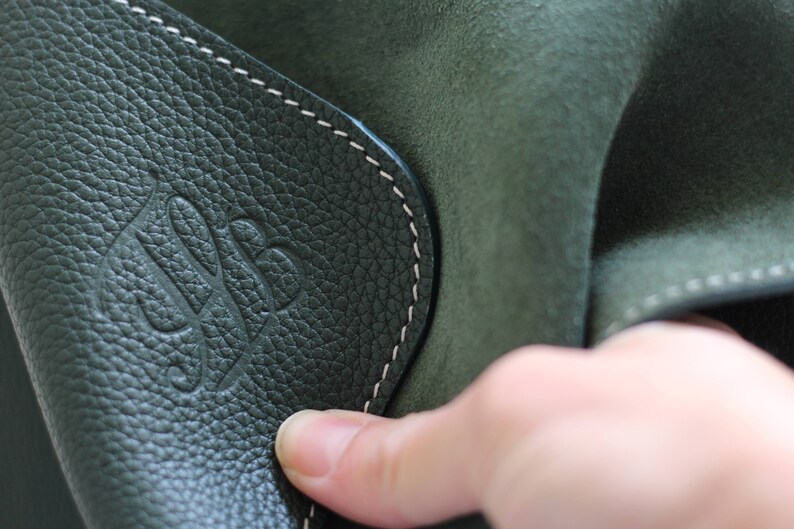 May include: Close-up of a dark green leather bag with a white stitched seam and a embossed logo. The logo is a stylized letter 'S' with a leaf design.