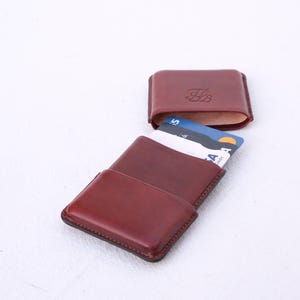 May include: A burgundy leather cardholder with a matching, open top case. The cardholder is open, revealing several credit cards. The top case has an embossed design. The items are on a white surface.