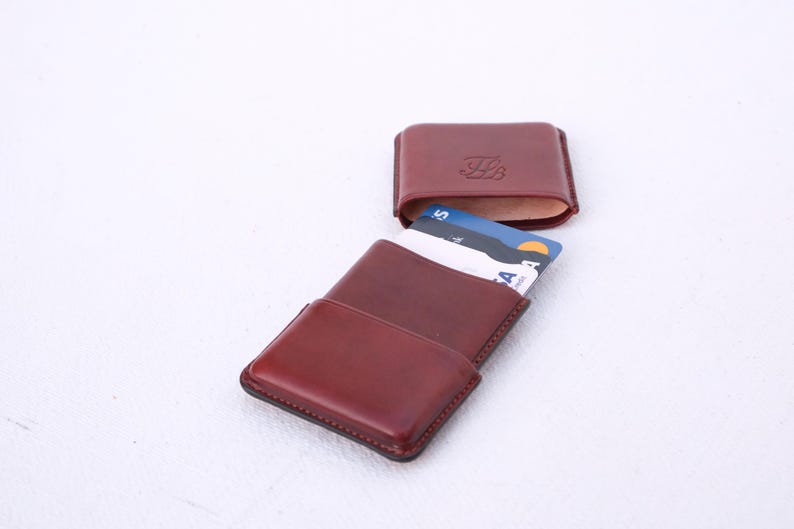 May include: A burgundy leather cardholder with a matching, open top case. The cardholder is open, revealing several credit cards. The top case has an embossed design. The items are on a white surface.