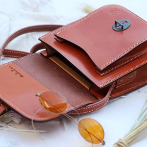 K&ouml;nnte beinhalten: Eine braune Leder-Umh&auml;ngetasche mit Klappenverschluss und einer Metallschnalle. Die Tasche hat ein kleines Fach auf der Vorderseite und ist ge&ouml;ffnet, um ein braunes Innenleben zu zeigen. Die Tasche liegt auf einer wei&szlig;en Oberfl&auml;che mit einem B&uuml;ndel getrockneter Weizenhalme.