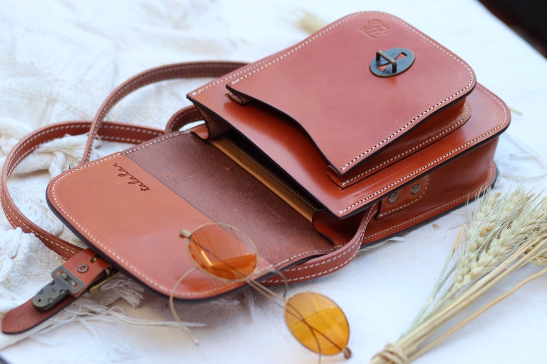 K&ouml;nnte beinhalten: Eine braune Leder-Umh&auml;ngetasche mit Klappenverschluss und einer Metallschnalle. Die Tasche hat ein kleines Fach auf der Vorderseite und ist ge&ouml;ffnet, um ein braunes Innenleben zu zeigen. Die Tasche liegt auf einer wei&szlig;en Oberfl&auml;che mit einem B&uuml;ndel getrockneter Weizenhalme.