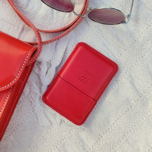 May include: A red leather case with a debossed monogram, next to a matching red crossbody bag and sunglasses. The case is rectangular and has a hinged lid. The bag has white stitching. The sunglasses have a thin silver frame and pink lenses.