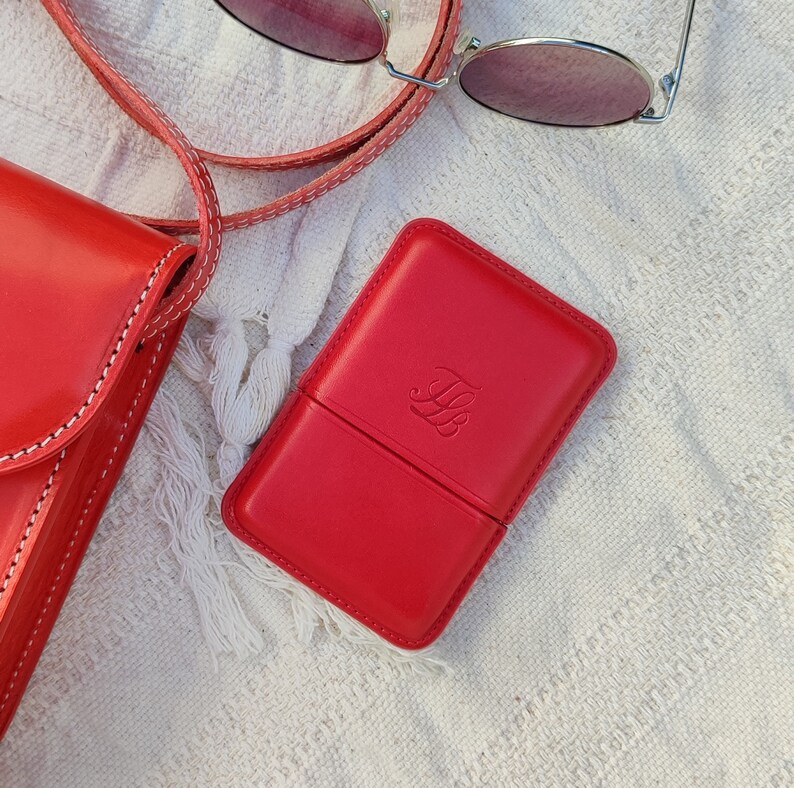 May include: A red leather case with a debossed monogram, next to a matching red crossbody bag and sunglasses. The case is rectangular and has a hinged lid. The bag has white stitching. The sunglasses have a thin silver frame and pink lenses.