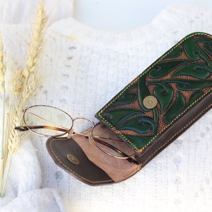 May include: A green leather glasses case with a floral design and brown stitching. The case is open and a pair of glasses is inside.