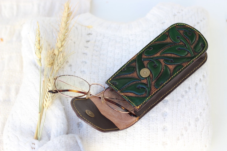 May include: A green leather glasses case with a floral design and brown stitching. The case is open and a pair of glasses is inside.
