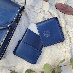 May include: A navy blue leather case with a white interior, a matching square card holder, and a navy blue crossbody bag with white stitching. The card holder has an embossed initial. A pair of sunglasses with pink lenses rests nearby.