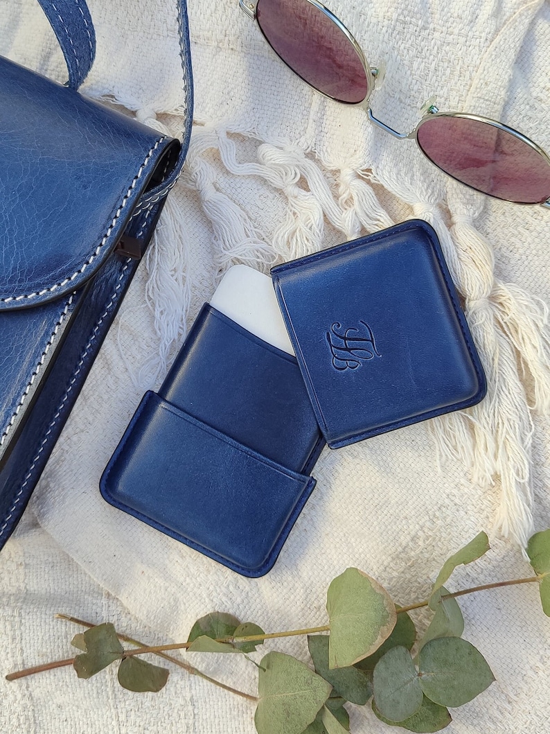 May include: A navy blue leather case with a white interior, a matching square card holder, and a navy blue crossbody bag with white stitching. The card holder has an embossed initial. A pair of sunglasses with pink lenses rests nearby.