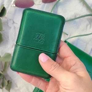 May include: A green leather glasses case with a debossed monogram. The case is rectangular with rounded corners and a hinged opening. Sunglasses with pink lenses are in the background.