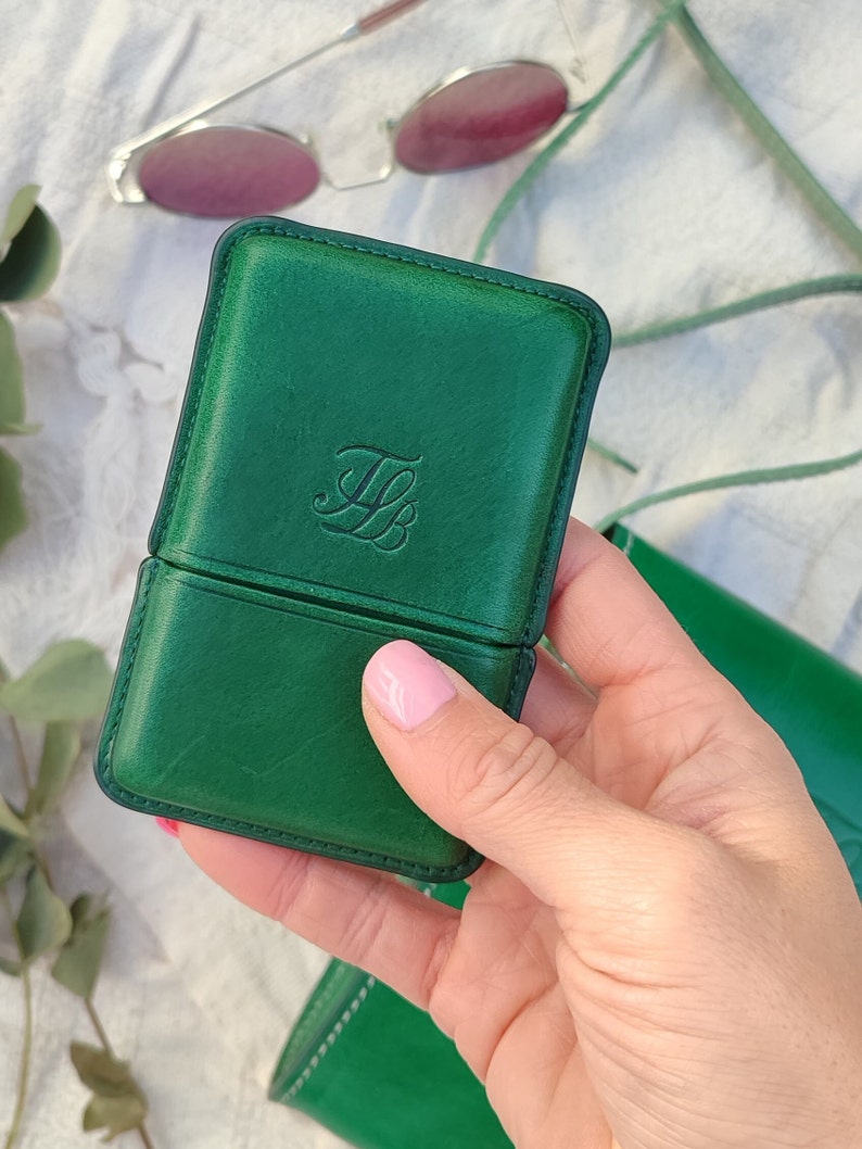 May include: A green leather glasses case with a debossed monogram. The case is rectangular with rounded corners and a hinged opening. Sunglasses with pink lenses are in the background.