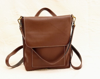 Puede incluir: Un bolso de cuero marrón cruzado con cierre de solapa y una sola correa. El bolso tiene un pequeño bolsillo en la parte delantera y un logotipo en relieve en la solapa.