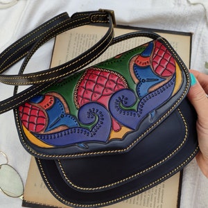 May include: Black leather crossbody bag with a colorful, hand-tooled design on the flap. The design features red, green, blue, and yellow floral and abstract patterns. The bag has a long, adjustable strap and gold stitching.