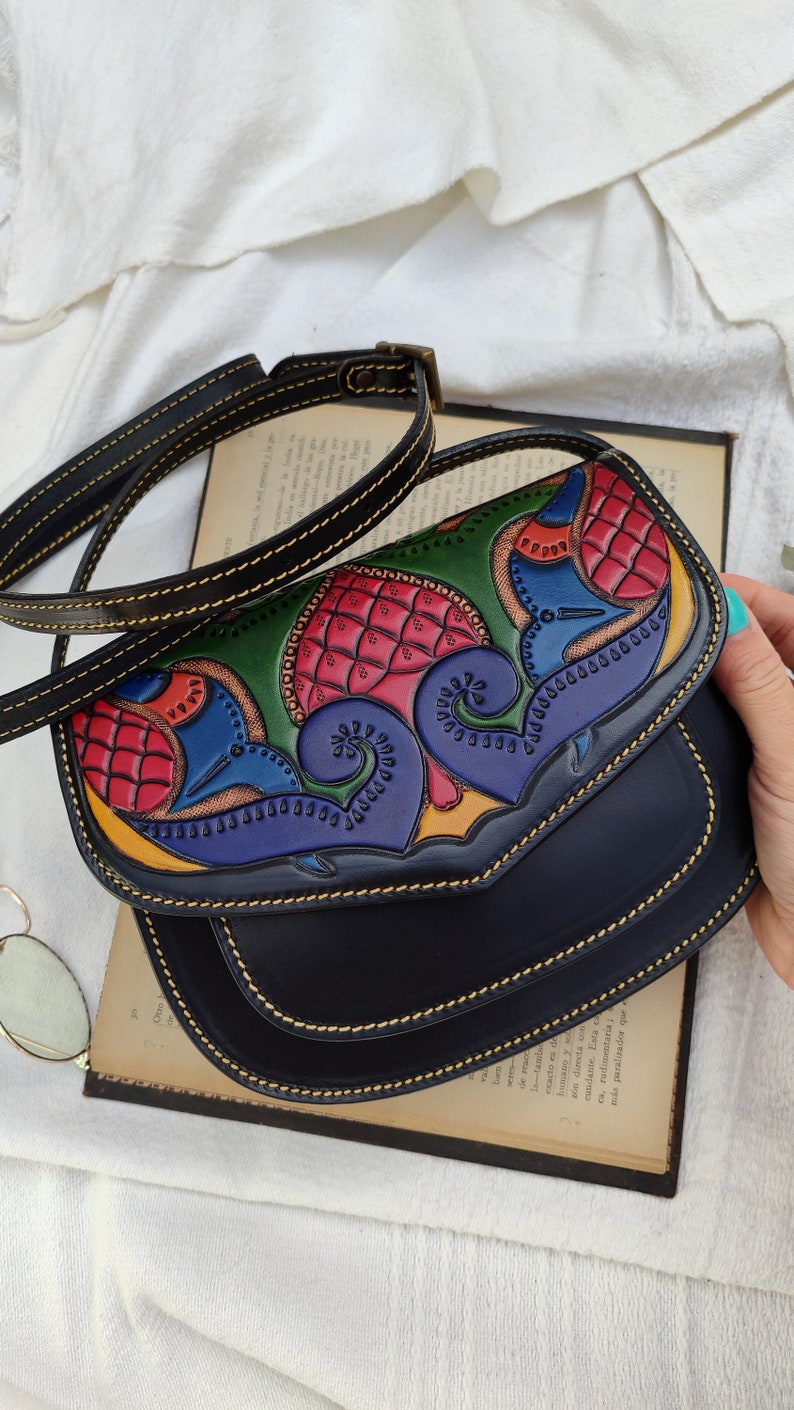 May include: Black leather crossbody bag with a colorful, hand-tooled design on the flap. The design features red, green, blue, and yellow floral and abstract patterns. The bag has a long, adjustable strap and gold stitching.