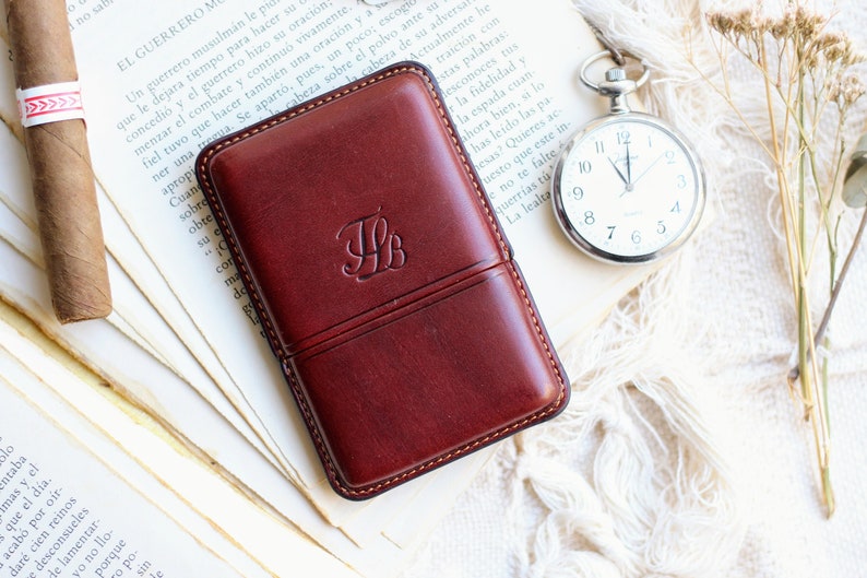 May include: A burgundy leather cigar case with an embossed monogram, a cigar, and a silver pocket watch on a vintage book page. The case has a stitched border.