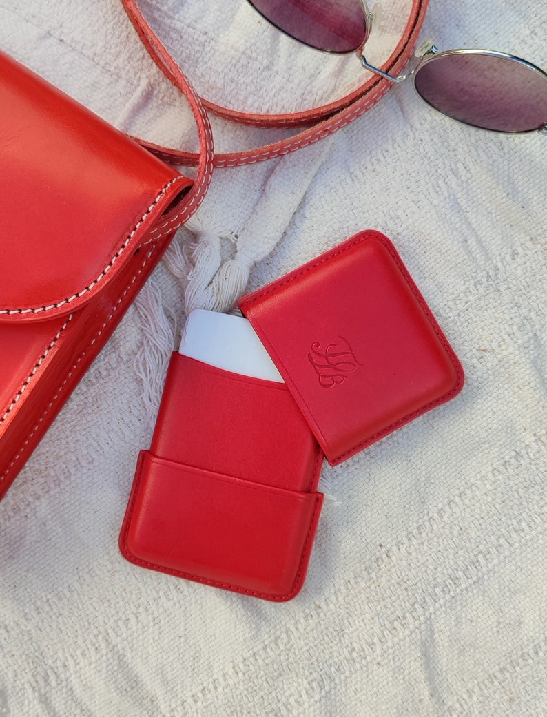 May include: Bright red leather crossbody bag and matching open case with a white interior. The case has an embossed monogram. A pair of round sunglasses with pink lenses rests nearby on a white textured surface.