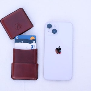 May include: A brown leather cardholder with cards, and a white smartphone with a black apple logo. The cardholder is open, revealing several credit cards. The cardholder is next to a closed, matching leather case.