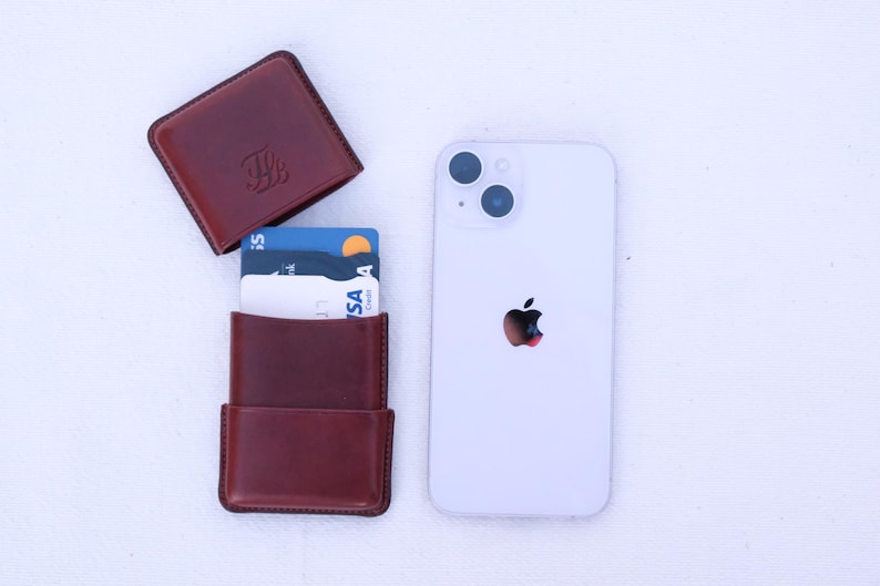 May include: A brown leather cardholder with cards, and a white smartphone with a black apple logo. The cardholder is open, revealing several credit cards. The cardholder is next to a closed, matching leather case.