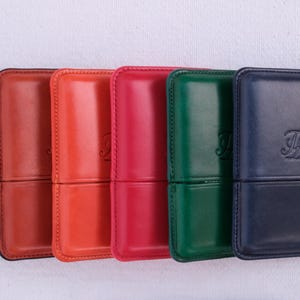 May include: A collection of six leather cases in various colors: burgundy, orange, red, green, and navy blue. Each case has a rectangular shape with rounded corners and a stitched seam. The cases have a debossed monogram on the front.