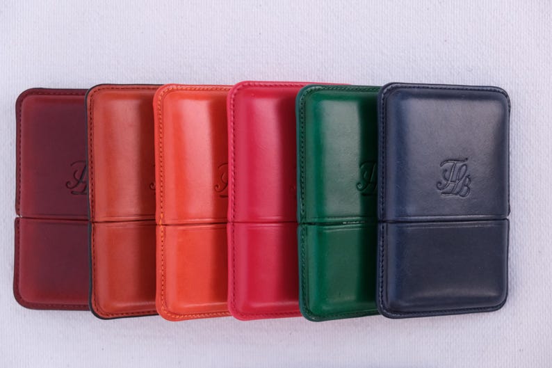 May include: A collection of six leather cases in various colors: burgundy, orange, red, green, and navy blue. Each case has a rectangular shape with rounded corners and a stitched seam. The cases have a debossed monogram on the front.