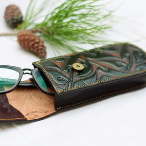 May include: A black leather glasses case with a green floral design and a brass button closure. The case is open and a pair of black glasses are inside.