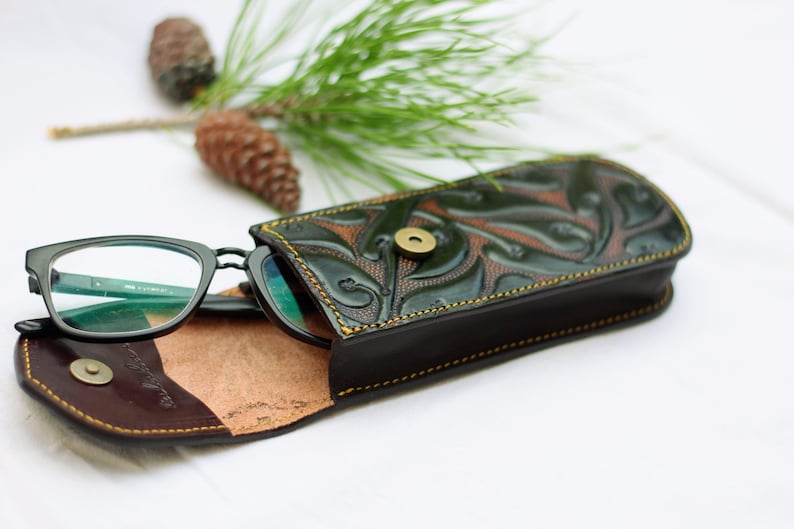 May include: A black leather glasses case with a green floral design and a brass button closure. The case is open and a pair of black glasses are inside.