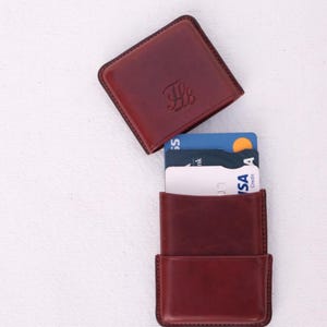 May include: A burgundy leather cardholder with a matching square top. The cardholder is open, revealing several credit cards. The top has an embossed design. The items are on a white surface.