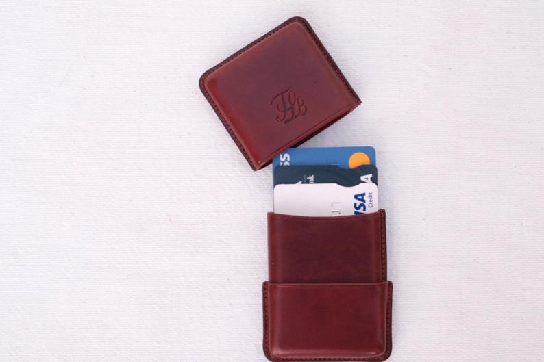 May include: A burgundy leather cardholder with a matching square top. The cardholder is open, revealing several credit cards. The top has an embossed design. The items are on a white surface.