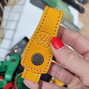 Puede incluir: Un llavero de cuero amarillo con un borde cosido y un anillo de metal. El llavero tiene una pequeña etiqueta de cuero redonda en relieve con el texto "TODDY" y un logotipo. El llavero también tiene las iniciales "LB" en relieve en el cuero.
