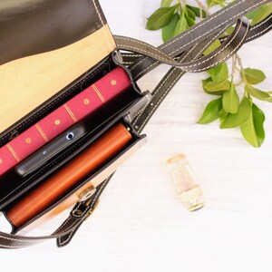 May include: A black and tan leather crossbody bag with a flap closure, open to reveal a red book, a black smartphone, and a brown leather wallet. The bag has a long strap with white stitching.