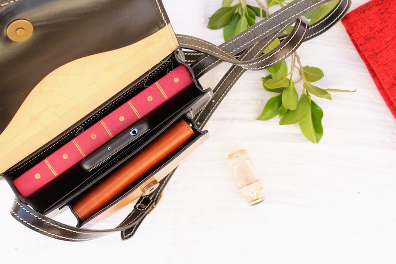 May include: A black and tan leather crossbody bag with a flap closure, open to reveal a red book, a black smartphone, and a brown leather wallet. The bag has a long strap with white stitching.
