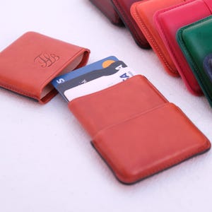 May include: Leather card holders in various colors, including orange, red, green, and navy blue. One orange holder is open, displaying credit cards. Another orange holder has initials embossed on the front. The holders are arranged on a white surface.
