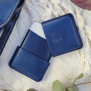 May include: A navy blue leather case, open to reveal a white object, with the initials "LB" embossed on the cover. The case is rectangular with rounded corners. A matching navy blue leather item is visible in the background.