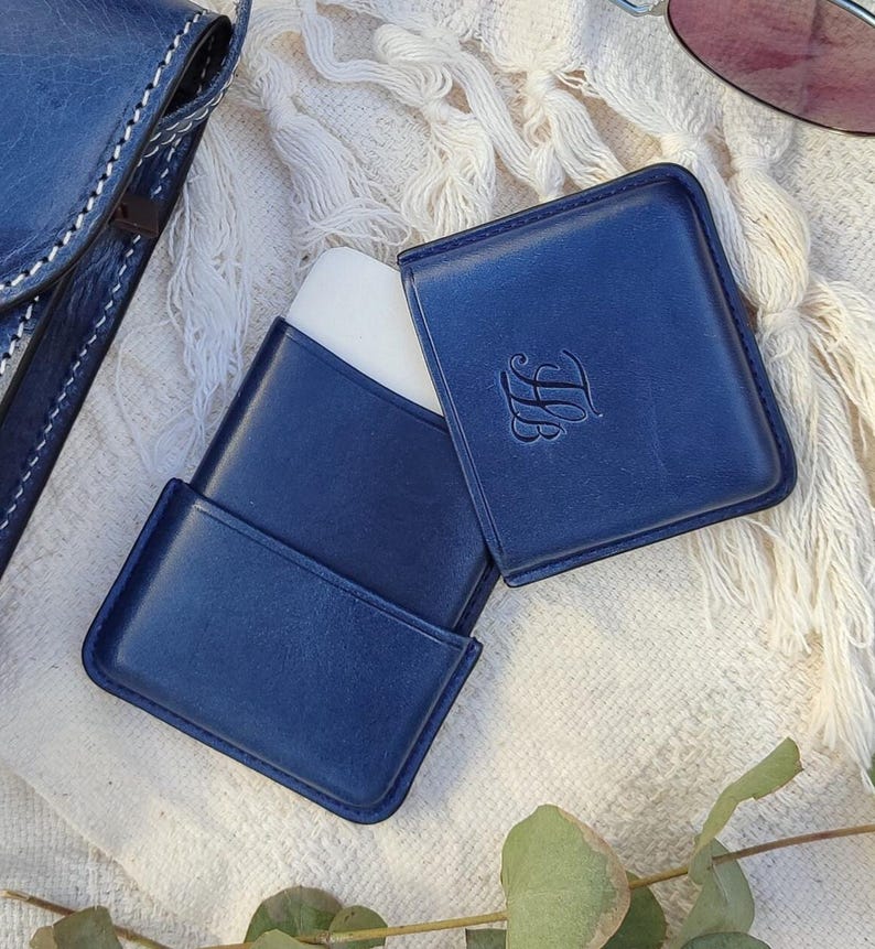 May include: A navy blue leather case, open to reveal a white object, with the initials "LB" embossed on the cover. The case is rectangular with rounded corners. A matching navy blue leather item is visible in the background.