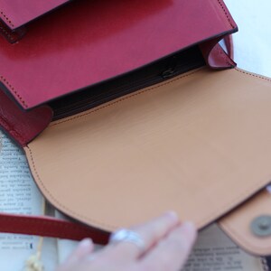 May include: A red leather satchel bag with a flap closure and a zippered pocket. The bag is open, revealing a tan leather interior.
