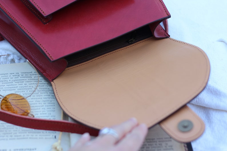 May include: A red leather satchel bag with a flap closure and a zippered pocket. The bag is open, revealing a tan leather interior.