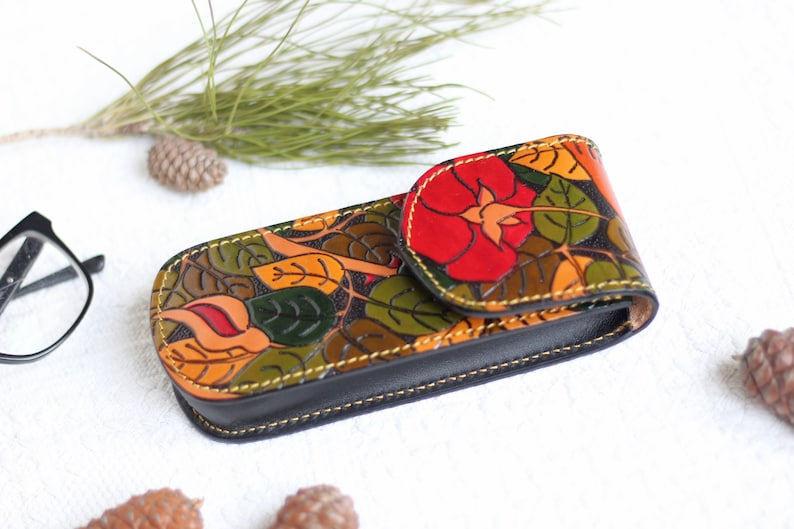 Op de afbeelding: Een bloemenpatroon lederen bril etui met een rood bloemenontwerp. De etui is gesloten en heeft een gele stiksel rond de randen.
