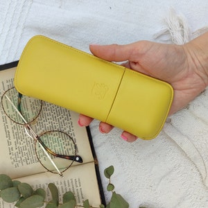 yellow leather glasses case with talabar logo engraved on it, a woman's hand holds the glasses case under an old open book