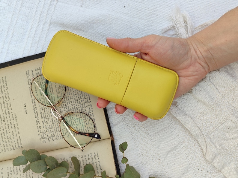 yellow leather glasses case with talabar logo engraved on it, a woman's hand holds the glasses case under an old open book