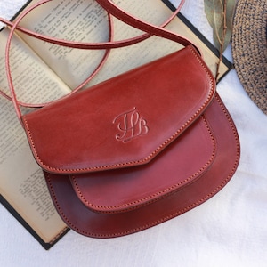May include: A burgundy leather crossbody bag with a flap closure and a monogrammed initial "HB" on the front.