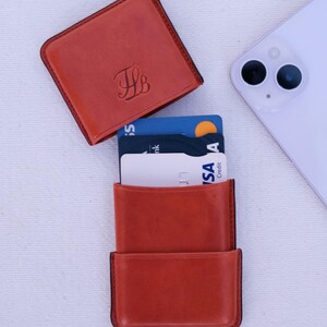May include: A burnt orange leather cardholder with a matching square top. The cardholder is open, revealing several credit cards. A white smartphone is placed to the side. The top of the cardholder has the initials "JB" in a script font.