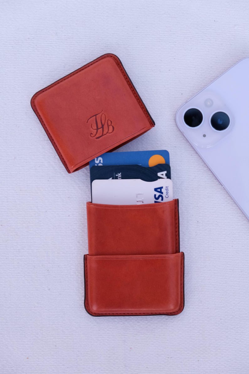 May include: A burnt orange leather cardholder with a matching square top. The cardholder is open, revealing several credit cards. A white smartphone is placed to the side. The top of the cardholder has the initials "JB" in a script font.