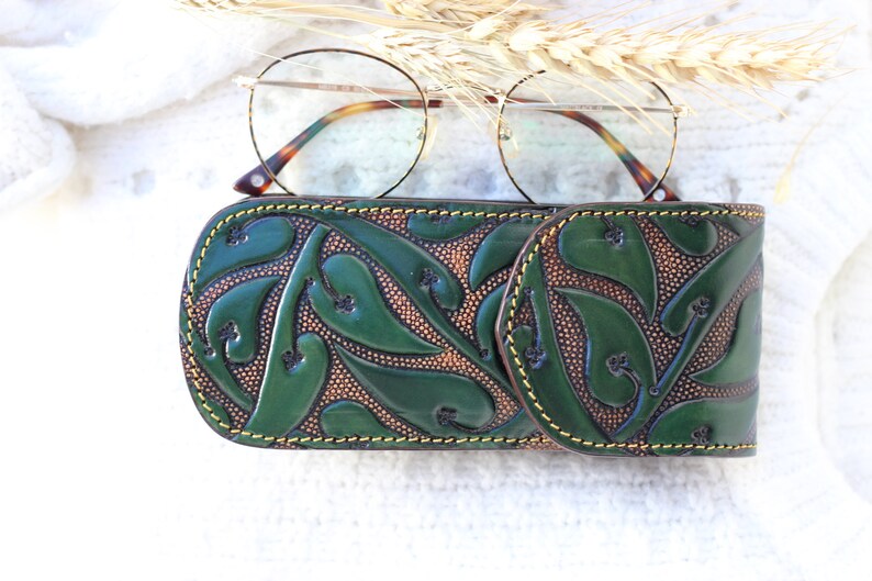 May include: A green leather glasses case with a floral pattern. The case has a yellow stitch around the edges.