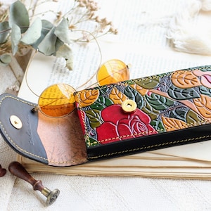 Op de afbeelding: Een bloemenpatroon lederen brilletui met een rood rozenontwerp. De etui is open en een paar ronde, oranje getinte brillen zitten erin.