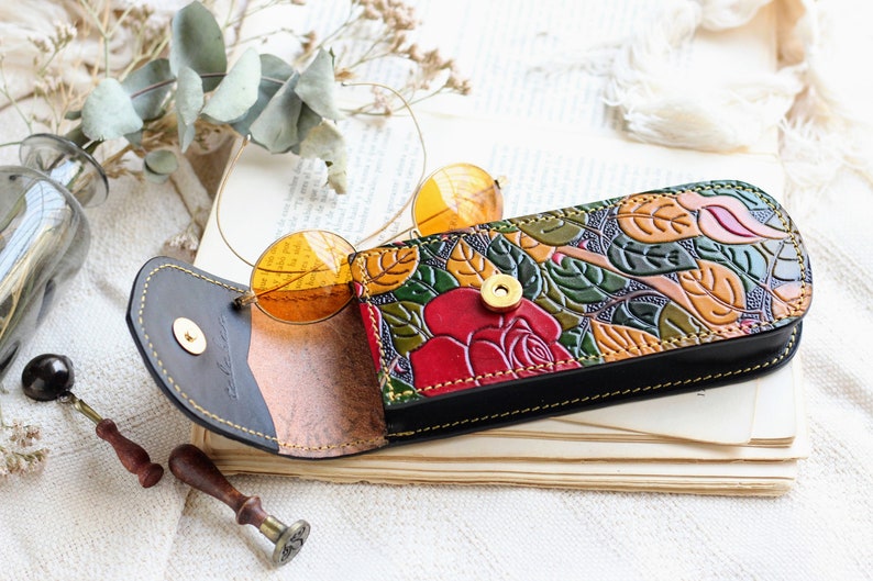 Op de afbeelding: Een bloemenpatroon lederen brilletui met een rood rozenontwerp. De etui is open en een paar ronde, oranje getinte brillen zitten erin.