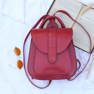 May include: A red leather backpack with a flap closure and a small pocket on the front. The backpack has a top handle and adjustable straps.