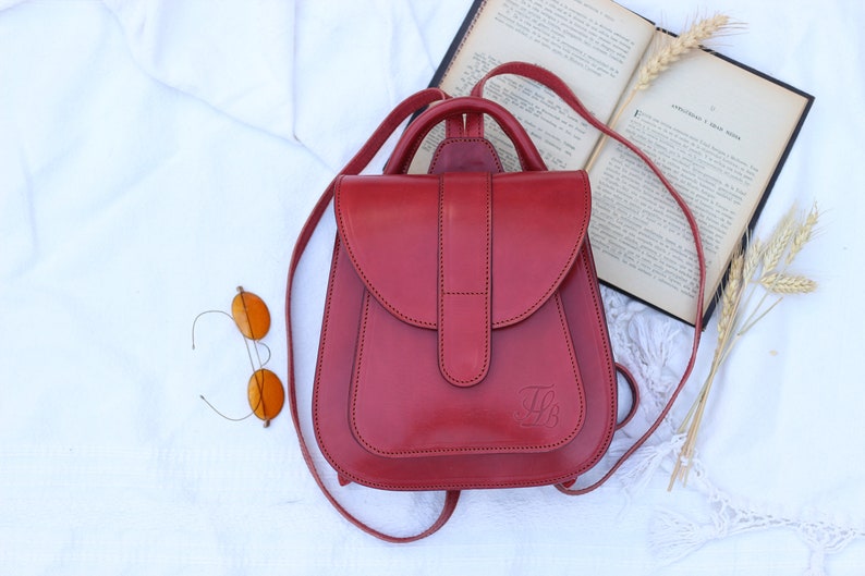 May include: A red leather backpack with a flap closure and a small pocket on the front. The backpack has a top handle and adjustable straps.