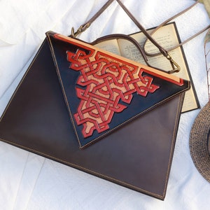 Könnte beinhalten: Eine braune Ledertasche mit Klappenverschluss und einem dekorativen roten und braunen Lederdesign. Die Tasche hat einen langen Riemen und einen oberen Griff.