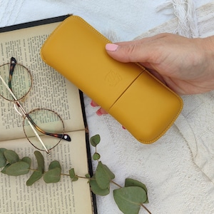 mustard  leather glasses case with talabar logo engraved on it, a woman's hand holds the glasses case under an old open book