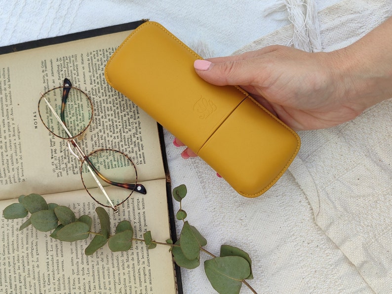 mustard  leather glasses case with talabar logo engraved on it, a woman's hand holds the glasses case under an old open book