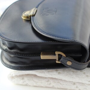 Puede incluir: Un bolso de cuero negro con cierre de lat&oacute;n y una correa decorativa. El bolso tiene cierre de solapa y asa superior.