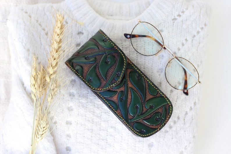 May include: A green leather glasses case with a floral design and yellow stitching. The case is closed and lying on a white knitted surface.